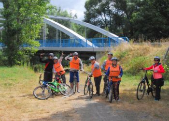 Powiększ zdjęcie: podopieczni w pomarańczowych kamizelkach i terapeuci z rowerami na tle lasu i mostu