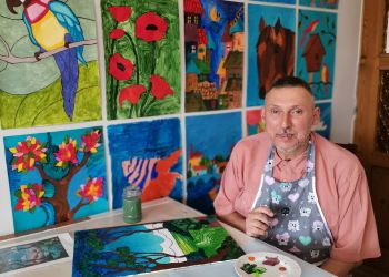 Powiększ zdjęcie: mężczyzna przy stole maluje obrazek, w tle na ścianie kolorowe obrazki