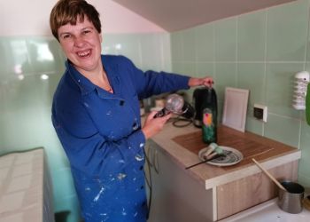 Powiększ zdjęcie: kobieta w niebieskim fartuchu stoi przy stole i suszy suszarką pomalowaną butelkę