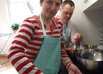 Powiększ zdjęcie: kobieta w bluzce w biało-czerwone paski i zielonym fartuszku kuchennym stoi przy blacie kuchennym i kroi masło, w tle mężczyzna, przed nimi dwie blaszane miski