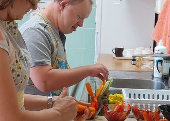 Powiększ zdjęcie: kobieta i mężczyzna przygotowują przy blacie kuchennym posiłek, do salaterek wkładają pokrojone w paski kolorowe warzywa: marchewki, papryki, ogórki oraz rzodkiewki