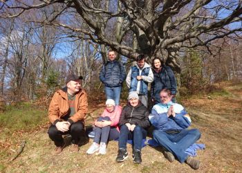 Powiększ zdjęcie: grupa siedmiu osób na tle drzewa z rozłozystymi konarami bez liści, cztery osoby z przodu siedzą, trzy osoby stoją za nimi