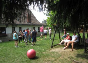 Powiększ zdjęcie: podczas zajęć w ogrodzie na pierwszym planie duża czerwona piłka i dwie osoby siedzące na drewnianej chuśtawce, grupa kobiet i mężczyzn widoczna z tyłu podczas ćwiczeń na ławeczce gimnastycznej