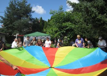 Powiększ zdjęcie: grupa około dziesięciu osób stoi w ogrodzie i trzyma przed sobą rozwiniętą chustę w kolorach tęczy