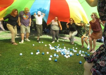 Powiększ zdjęcie: grupa siedmiu osób stoi pod tęczową chustą podtrzymując ją, przed nimi na trawie leżą kolorowe, plastikowe piłeczki