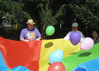 Powiększ zdjęcie: dwóch mężczyzn i kobieta trzymają krawędzie chusty w kolorach czerwonym, żółtym, zielonym i niebieskim, nad chustą unosi się pięc kolorowych balonów