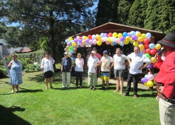 Powiększ zdjęcie: grupa ośmiu osób stoi na trawie w ogrodzie, każdy trzyma się za biodra, na pierwszym planie mężczyzna w stroju góralskim i czarnym kapeluszu mówi do mikrofonu, za grupą w tle dach altany ustrojony kolorowymi balonami