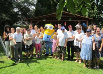 Powiększ zdjęcie: grupa około dwudziestu kobiet i mężczyzn stoi w ogrodzie, w środku pomiędzy nimi stoi minionek, żółta postać w niebieskich spodniach i okularach