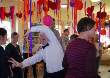 Powiększ zdjęcie: grupa osób tańczy w sali ustrojonej w wiszące fioletowo-różowe pasy z bibuły i balony