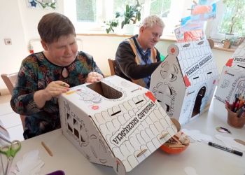 Powiększ zdjęcie: kobieta i mężczyzna siedzą przy stole i składają białe kartony w kształcie domków