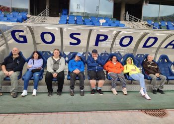 Powiększ zdjęcie: osiem osób siedzi na niebieskich fotelach na ławce zawodników na stadionie, nad nimi duże niebieskie litery na białym tle tworzą napis gospoda