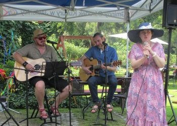 Powiększ zdjęcie: kobieta w różowej sukience i czarnym kapeluszu trzyma mikrofon i śpiewa, z tyłu dwaj mężczyźni siedzą i grają na gitarach