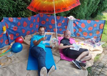 Powiększ zdjęcie: dwóch podopiecznych wyleguje się na zaaranżowanej w ogrodzie plaży, leżą na jasnym kocu, czytają gazety, pod parasolem, w tle kolorowy parawan