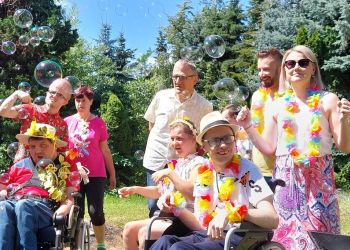 Powiększ zdjęcie: grupa kilku podopiecznych i terapeutów w kolorowych ubraniach i kwiatowych naszyjnikach, uśmiecha się, z przodu trzy osoby na wózkach