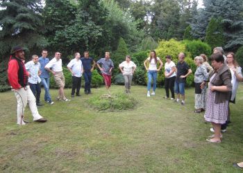 Powiększ zdjęcie: grupa około piętnastu podopiecznych, terapeutów i wodzireja w stroju góralskim stoi w półkolu w ogrodzie wykonując ruchy taneczne