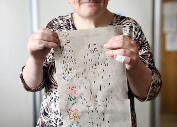 Powiększ zdjęcie: uczestniczka zajęć stoi i prezentuje wyszywaną pracę